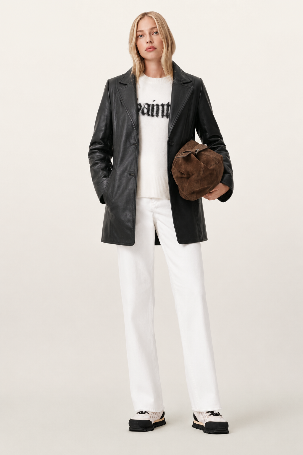 Woman in black leather jacket, white graphic sweater, white pants, and brown suede bag