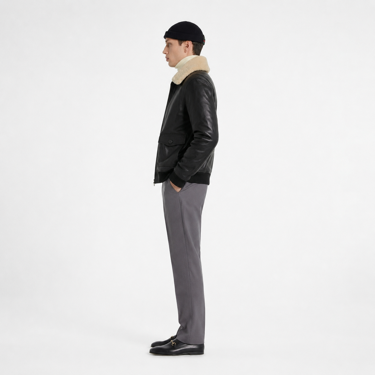 Man in black leather bomber jacket with shearling collar, gray trousers, black beanie, and loafers