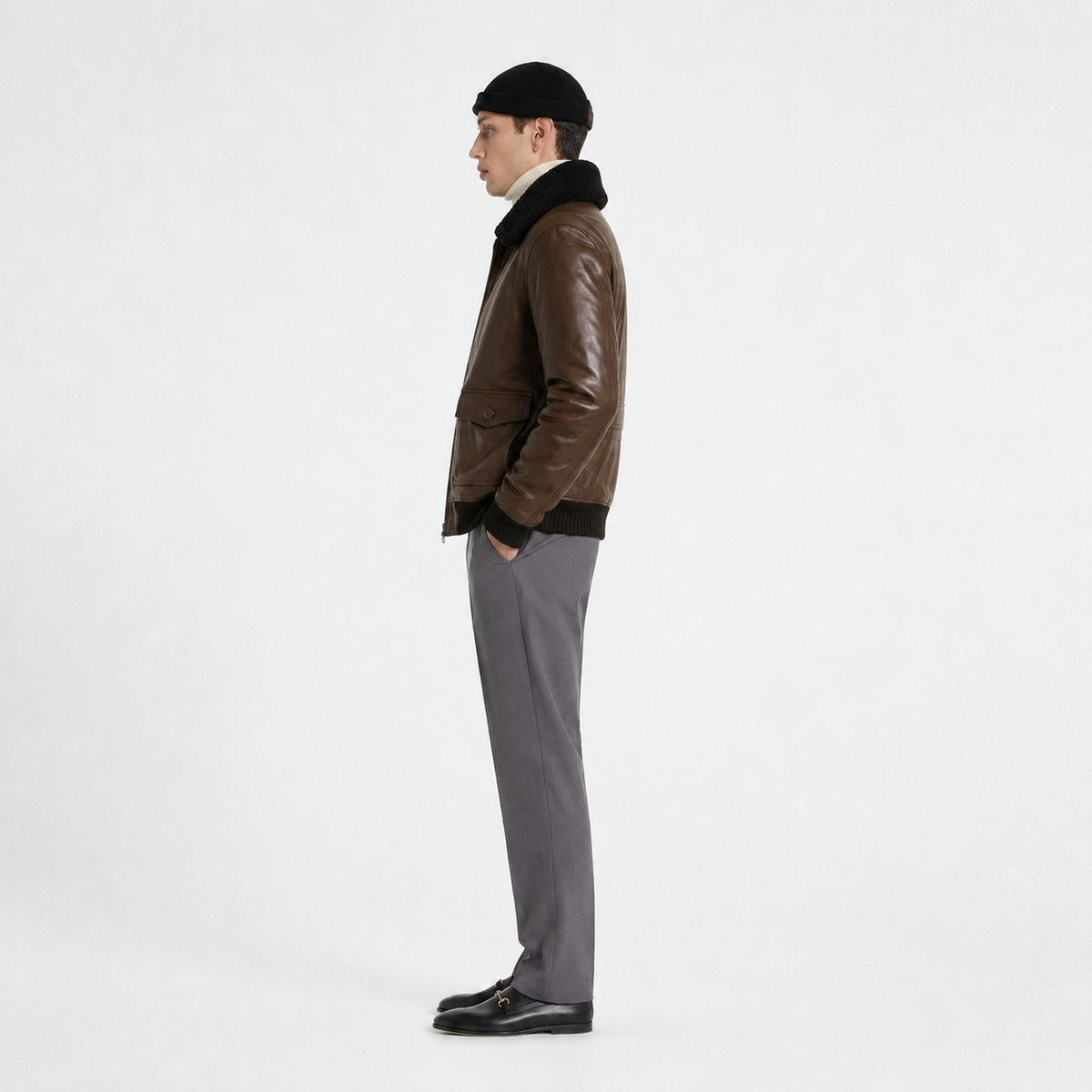Man wearing brown leather bomber jacket with shearling collar, gray trousers, black beanie, and loafers