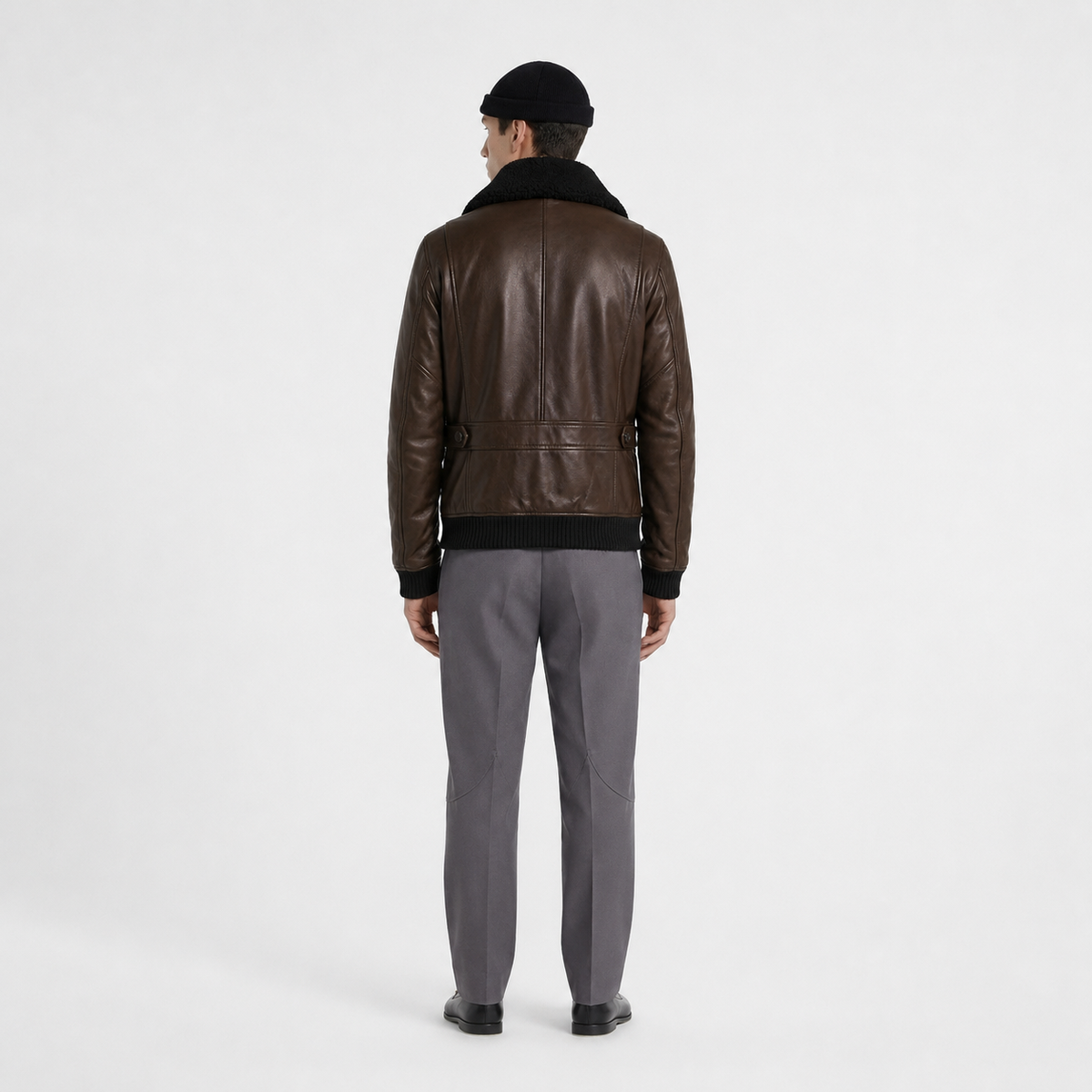 Men's brown leather bomber jacket with shearling collar, back view, paired with gray trousers and black beanie