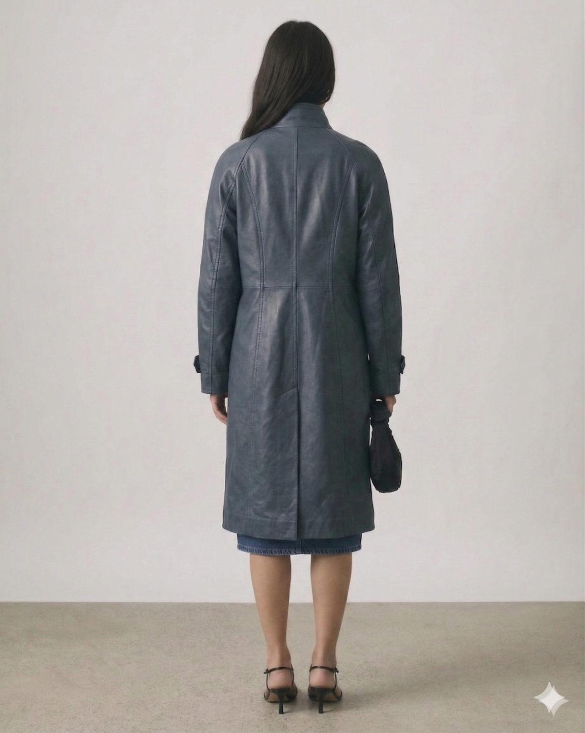 Woman wearing a long gray leather coat, denim skirt, and heels, holding a small black bag, back view