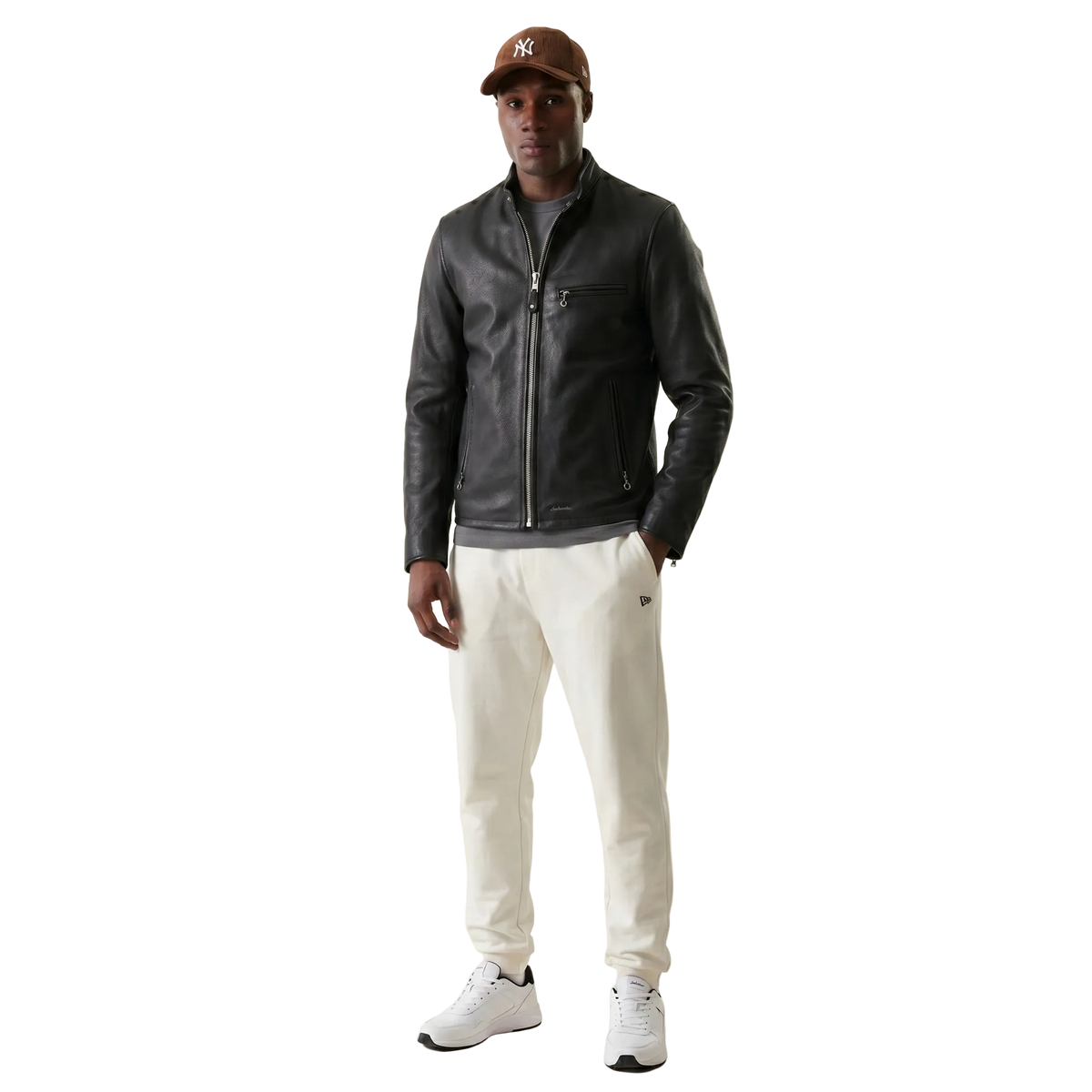 Men's black leather jacket with zipper, paired with cream joggers and white sneakers, styled with a brown cap.