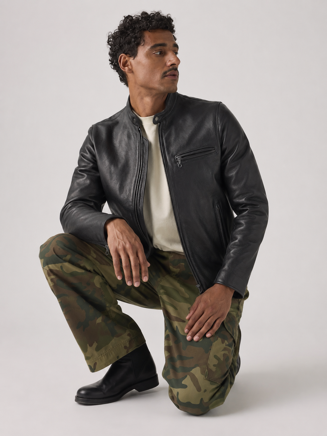 Man in black leather jacket, camo pants, and black boots, crouching against neutral background