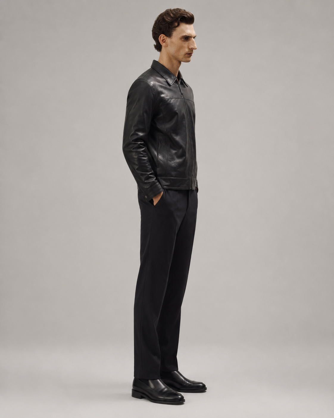 Man in black leather jacket, slim-fit trousers, and dress shoes, side profile on neutral background