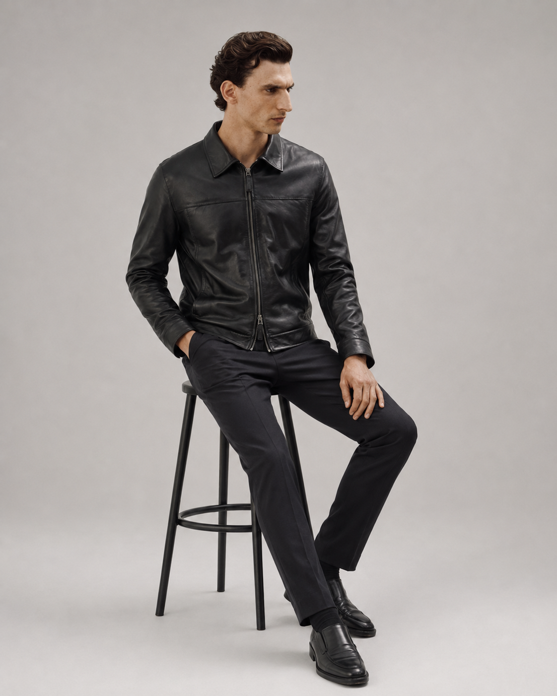 Man in black leather jacket and slim-fit trousers, seated on stool, modern men's fashion