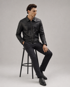 Man in black leather jacket and slim-fit trousers, seated on stool, modern men's fashion