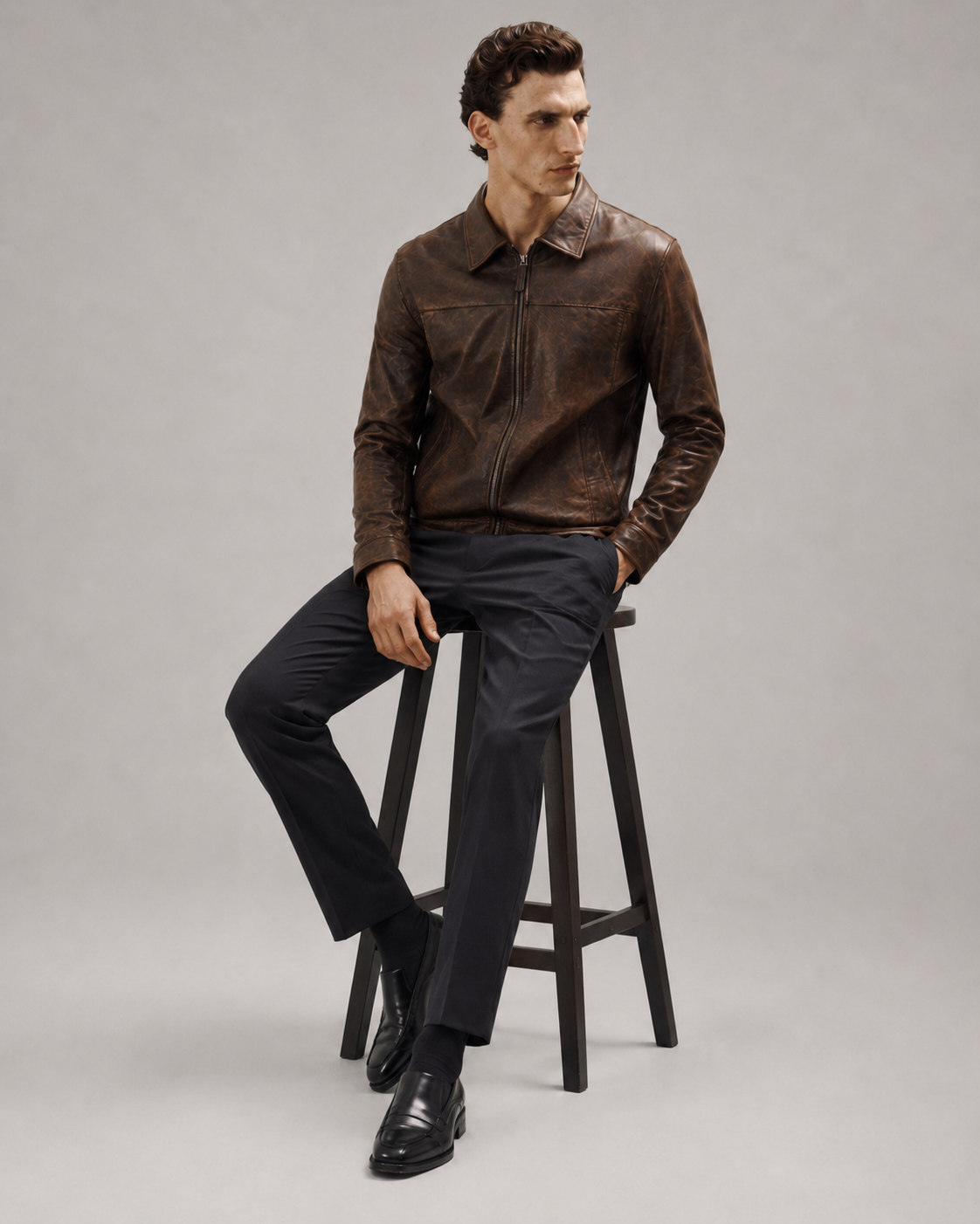 Man wearing brown leather jacket, black trousers, and loafers, seated on stool