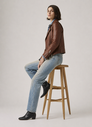 Woman in brown leather jacket, blue jeans, and black boots sitting on wooden stool