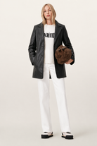 Woman in black leather jacket, white graphic sweater, white pants, and brown suede bag