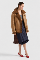 Women's brown shearling aviator jacket with plush collar, paired with navy pleated skirt and red heels