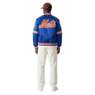 Blue Mets varsity jacket with orange lettering, worn by man in white pants and cap, back view