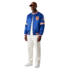 Men's royal blue New York Mets satin varsity jacket with orange trim and team logo, styled with white pants and cap
