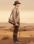 Man in desert wearing tan leather jacket, fedora hat, carrying satchel and coiled rope