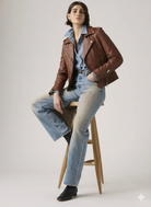 Woman in brown leather jacket, denim shirt, and wide-leg jeans seated on wooden stool