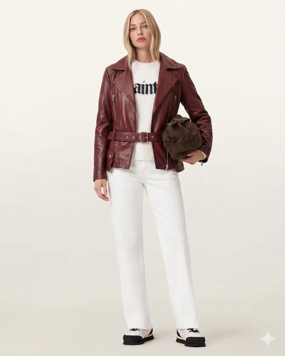 Woman in burgundy leather moto jacket, white jeans, and graphic sweater, holding brown bag