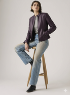 Woman in purple leather moto jacket, gray top, and blue jeans sitting on wooden stool