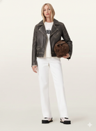 Woman in distressed leather moto jacket, white jeans, and chunky sneakers holding brown suede bag