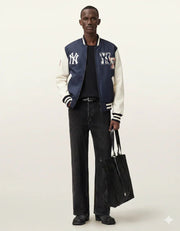 Man wearing navy and cream varsity jacket with NY logo, black jeans, and holding a tote bag