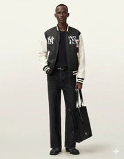 Man wearing black and white varsity jacket with NY logo, black jeans, holding tote bag